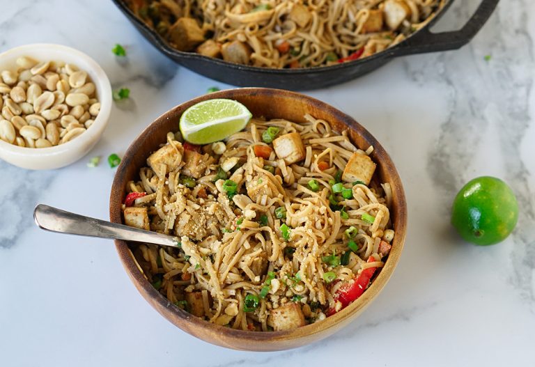 Bowl of vegan peanut butter pad Thai topped with crushed peanuts, green onions, and lime wedges